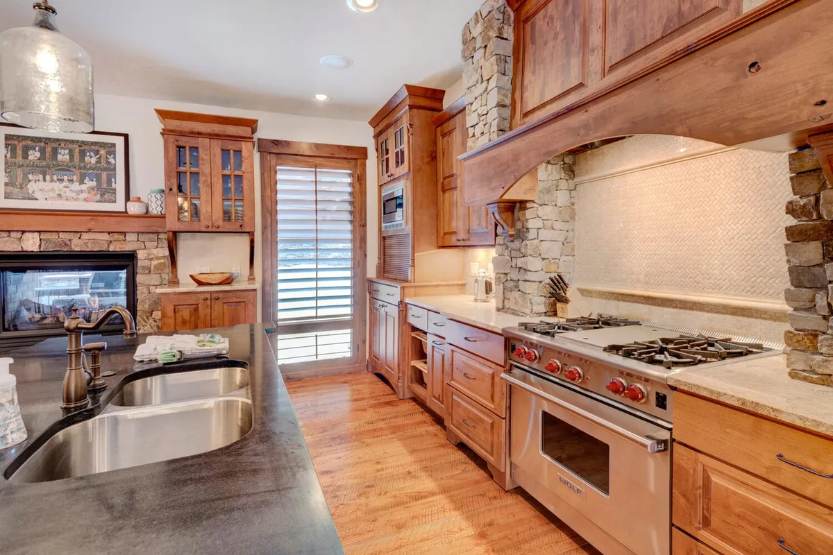 A cozy, open-plan kitchen and living area featuring a stone fireplace, wooden cabinetry, a kitchen island with sink, and stainless steel appliances. The setting feels welcoming, well-appointed, and perfect for both casual gatherings and festive occasions.