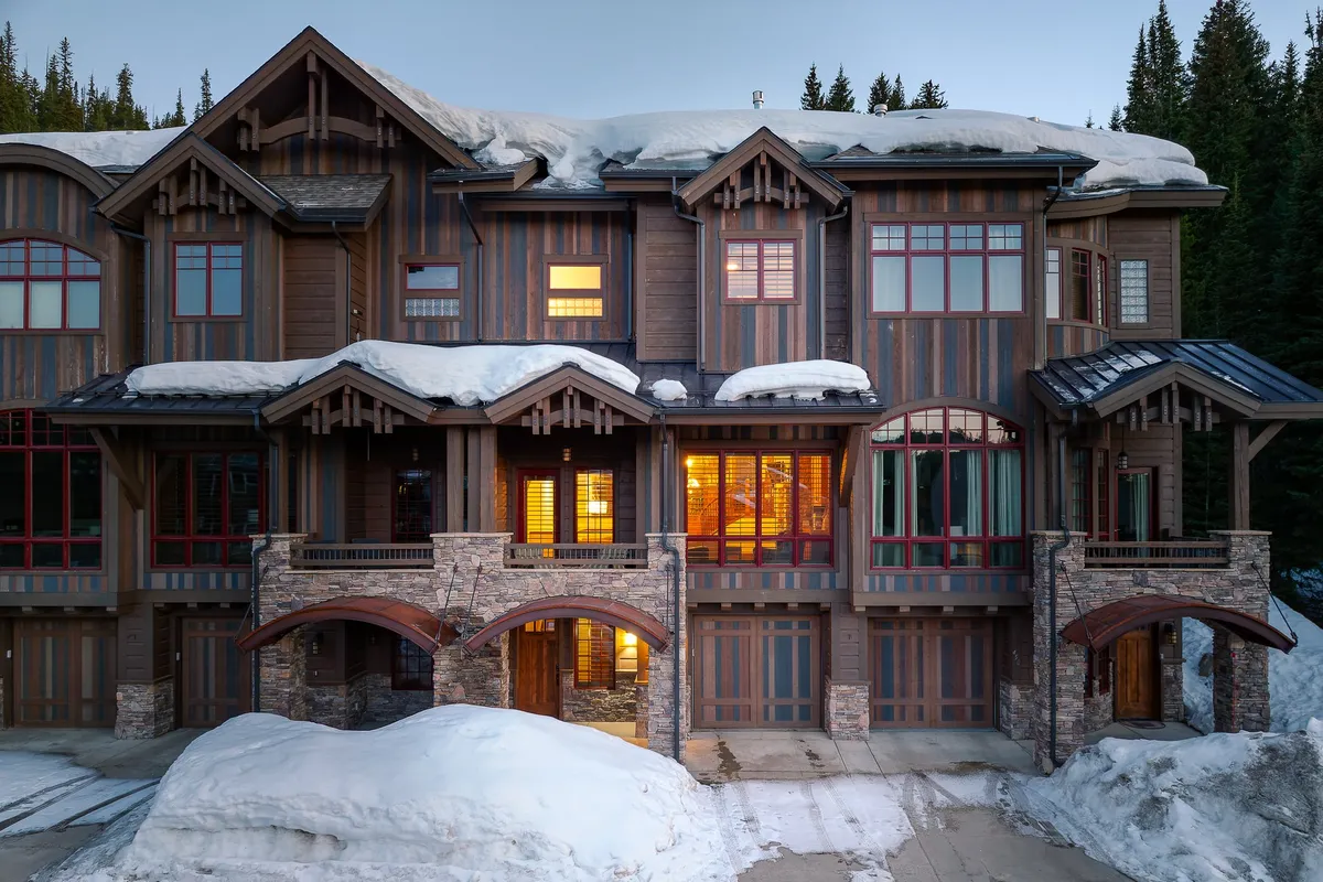 The image features the snow-covered exterior of a luxury villa in Winter Park, Colorado. The warm glow from the windows contrasts beautifully with the sparkling snow, while the stone façade and wooden beams evoke mountain charm and quality craftsmanship. The inviting porch and spacious yard hint at family-friendly outdoor activities and peaceful relaxation, all set against a serene, alpine backdrop.