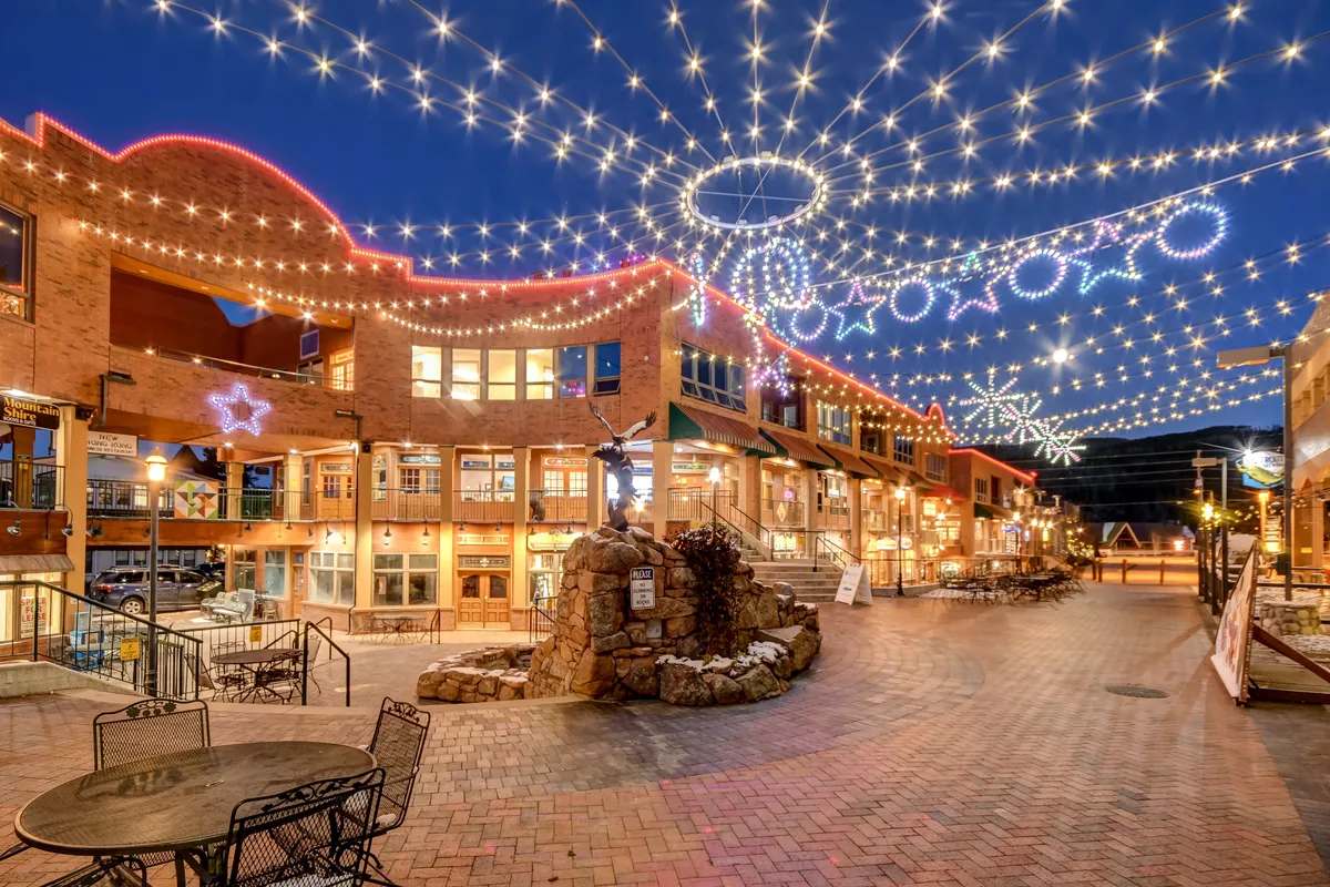 An inviting exterior shot of a community area with twinkling lights overhead, decorative stars and circles, and a central stone feature. People are seen dining, socializing, and enjoying an evening out. The scene feels festive and communal, perfect for families coming together after a day of adventure.