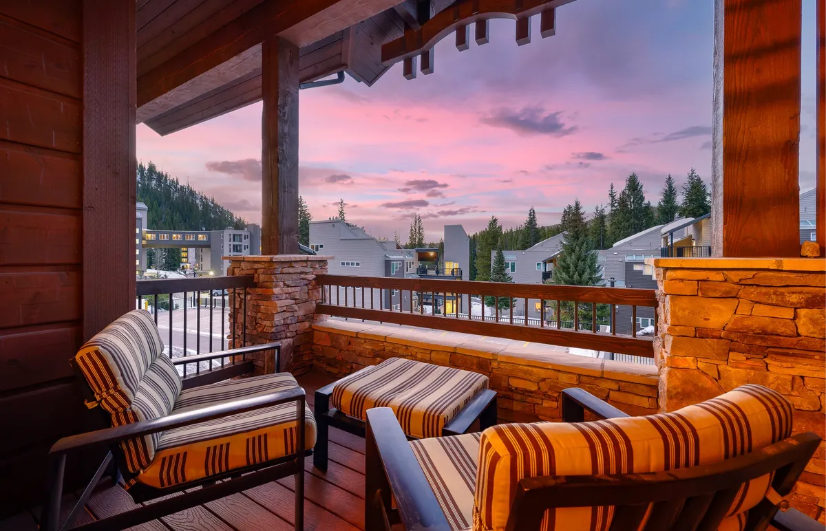 A tranquil patio setting with striped chairs overlooking a stunning sunset sky and mountain landscape, framed by wooden balcony railings. The scene evokes peaceful relaxation and connection to the natural surroundings.