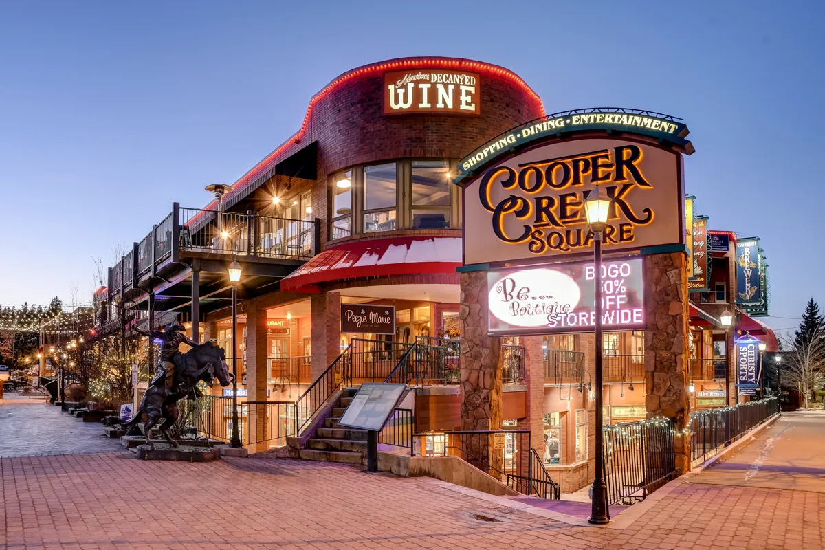 This exterior image captures the lively atmosphere of Cooper Creek Square, highlighting its vibrant signage, wine shop, distinctive architectural features, outdoor seating area, and a statue at the entrance. People are engaged in shopping, dining, and social activities against the backdrop of a welcoming plaza.