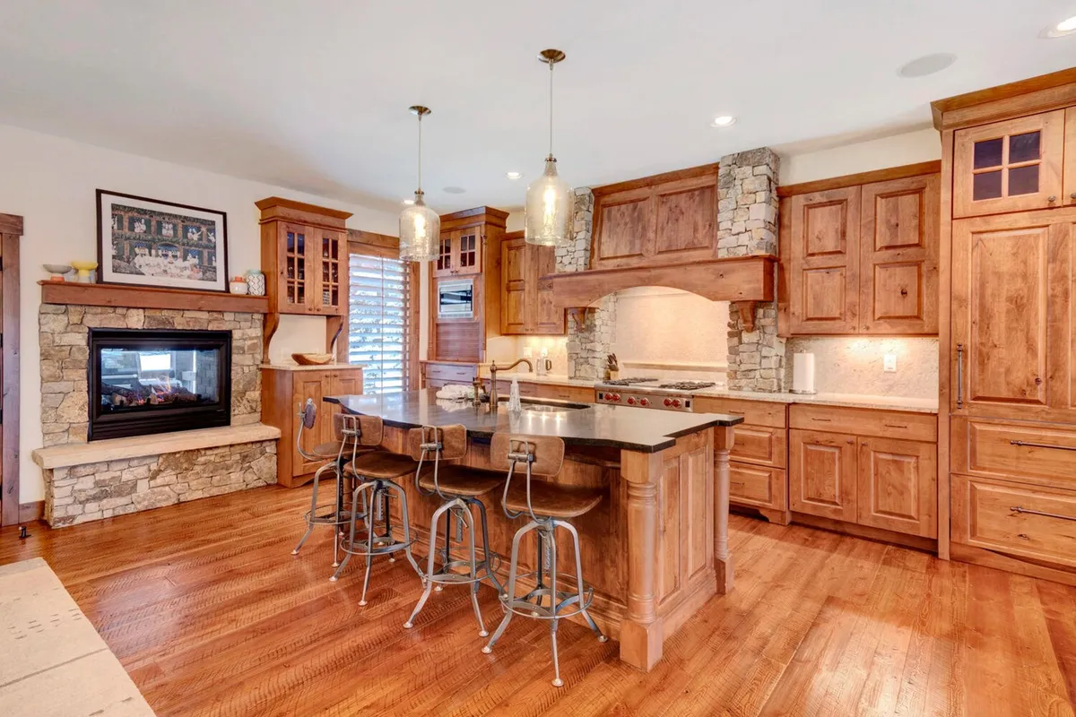 A warm, inviting kitchen featuring a prominent island with bar stools, wood cabinetry, and a crackling fireplace in the background. The kitchen is designed for both cooking and socializing, with open sightlines to the adjoining living area, and hints of premium appliances.