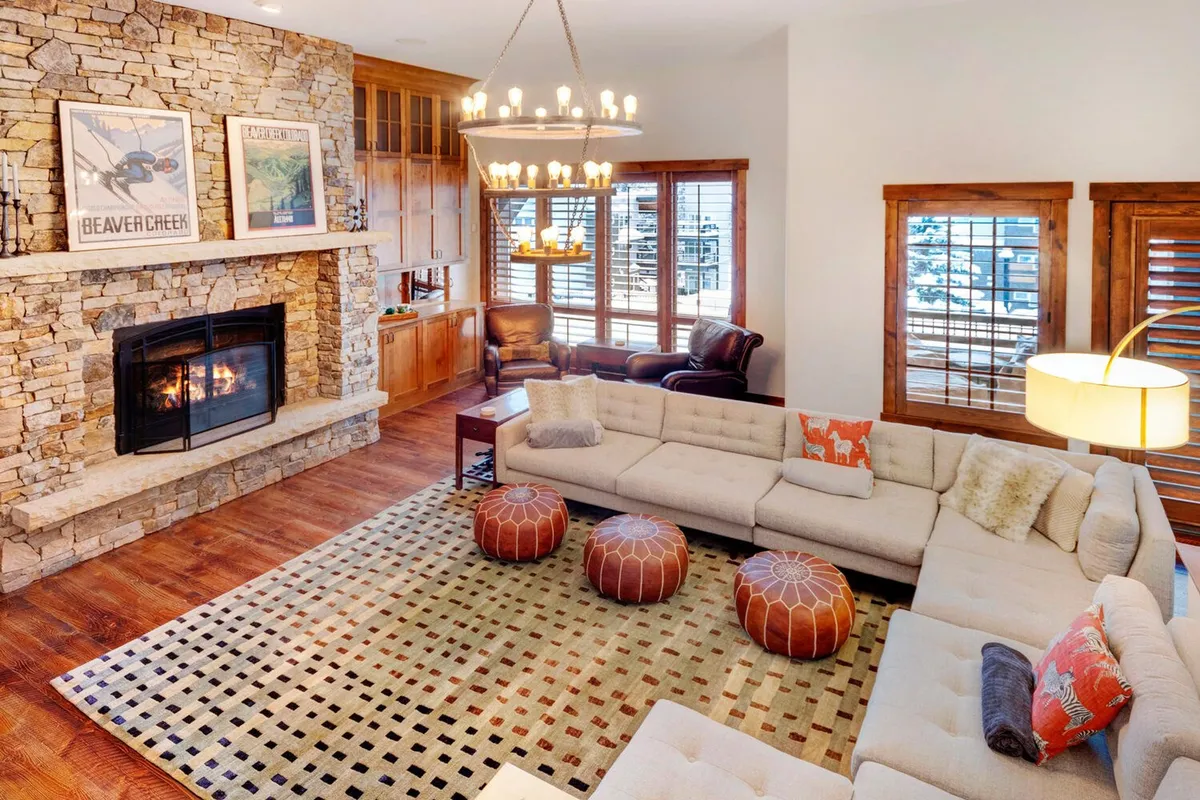A spacious open-concept living area featuring a majestic stone fireplace as the central focal point. Plush designer sectional sofa, colorful ottomans, and a striking chandelier set an inviting, cozy tone. Wooden shutters and large windows hint at natural light and scenic views, creating a warm and communal atmosphere.