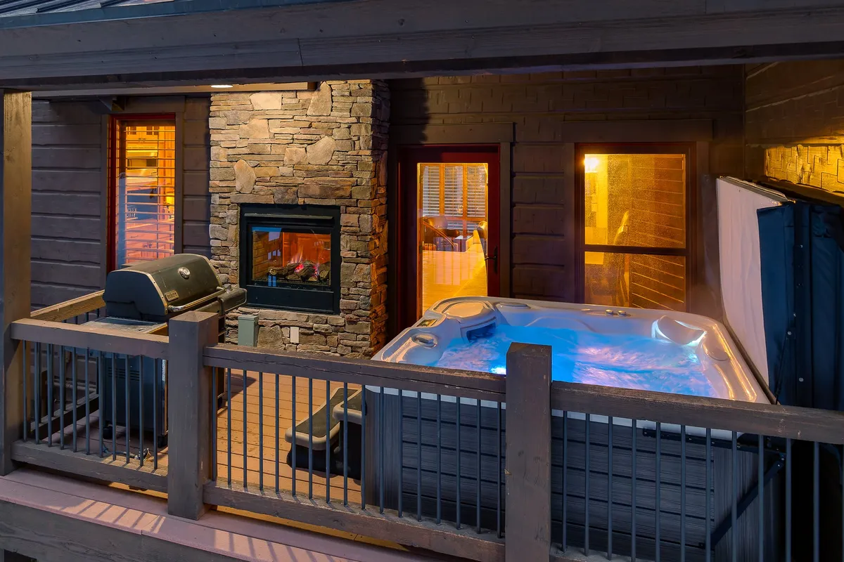 An inviting outdoor space featuring a private hot tub illuminated with LED lights, an outdoor fireplace, and a gas grill set against a mountain backdrop. The setup is perfect for relaxing evenings and social gatherings, blending luxury with alpine scenery.