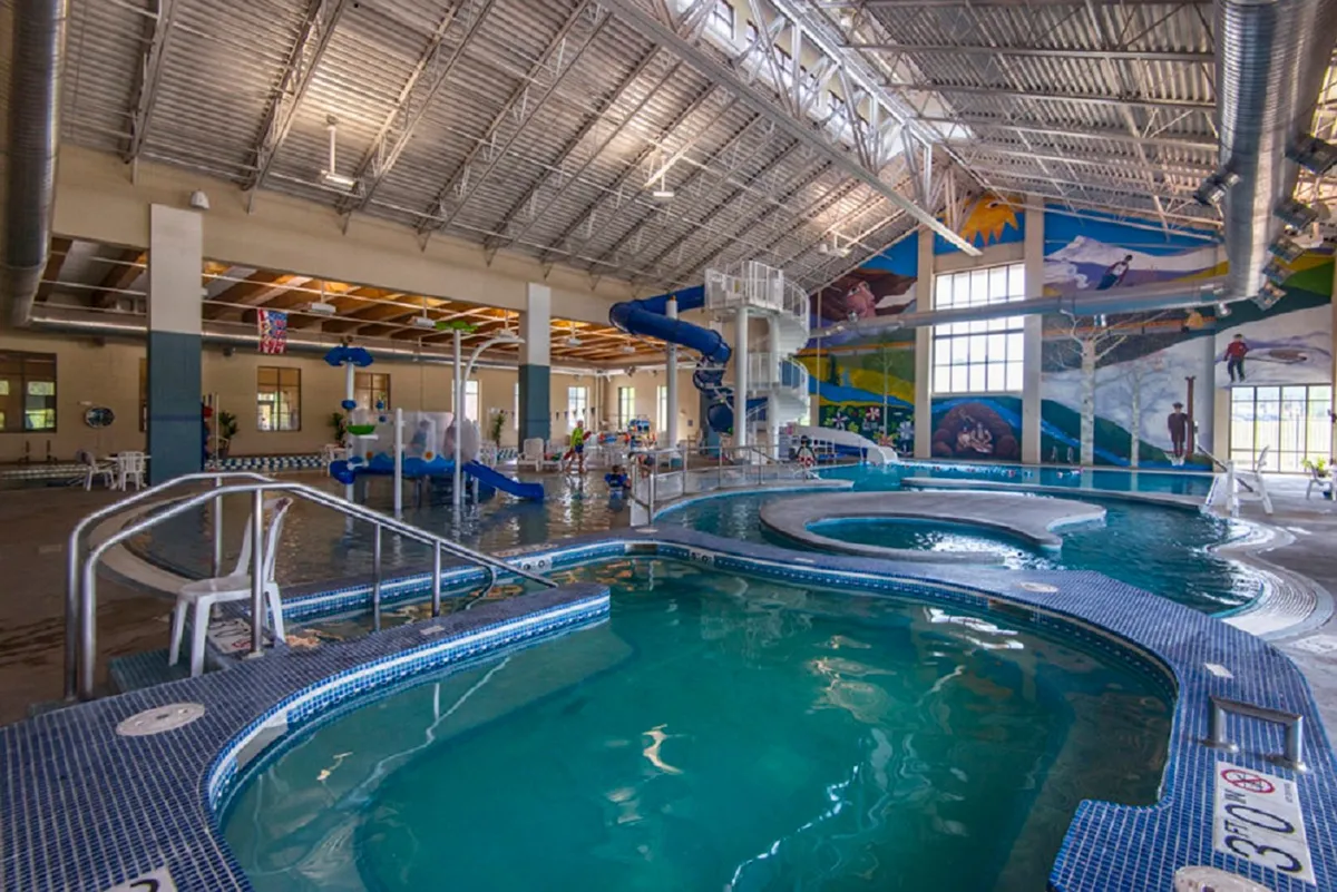 A lively indoor pool area with a water slide, colorful play structures, and a vibrant mural, bustling with energy and designed for family fun and children's activities.
