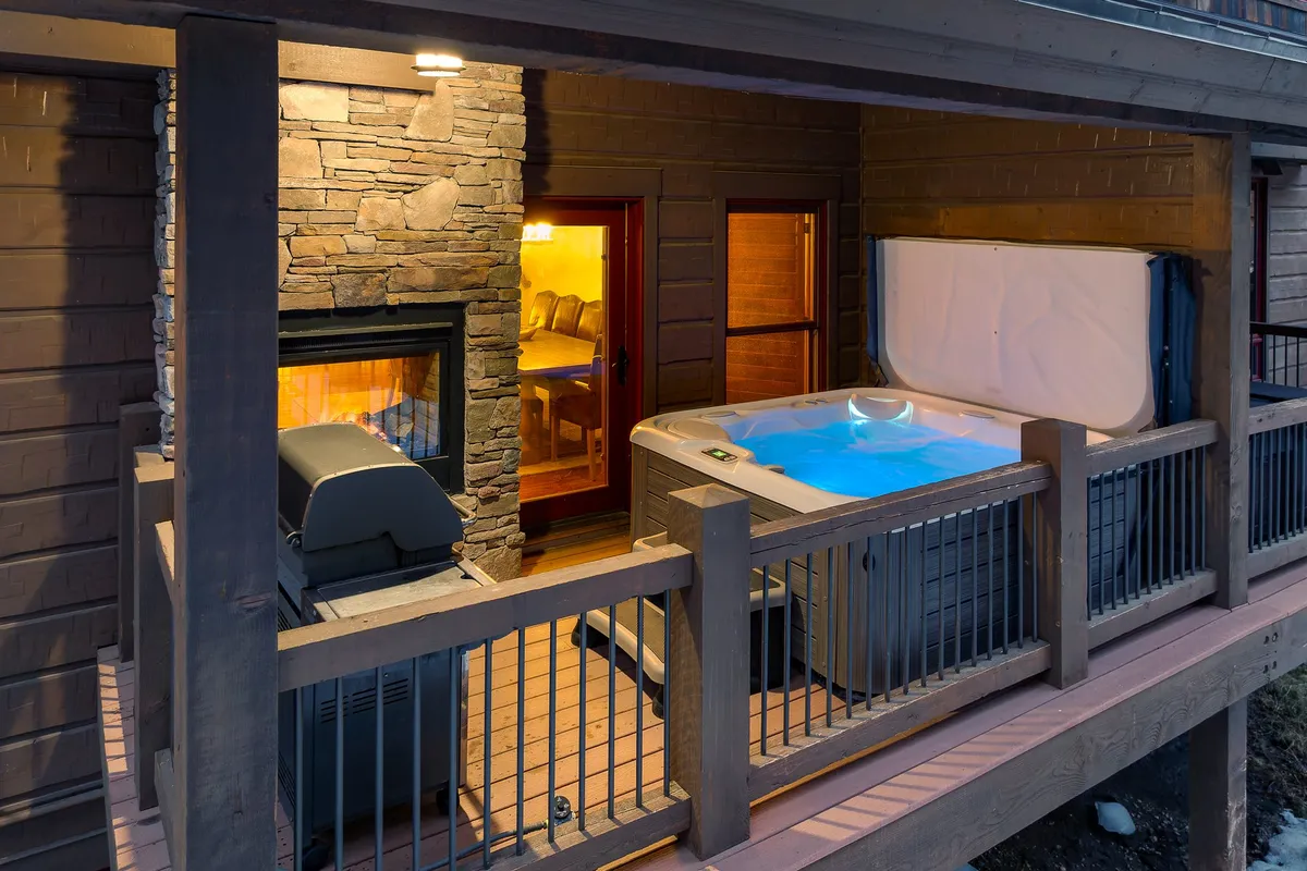 A spacious outdoor area featuring a private hot tub, stone fireplace, and a grill, with comfortable seating ideal for socializing or unwinding after a day of adventure. The ambiance is both warm and tranquil, enhanced by the natural mountain environment.