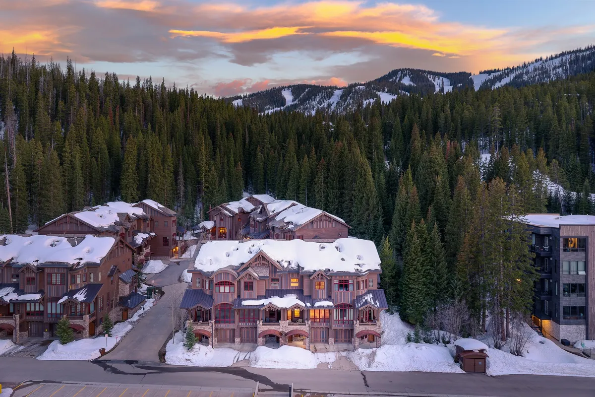 An inviting winter exterior scene featuring snow-laden roofs, tall pine trees, a dramatic mountain backdrop, and warmly lit windows, evoking the coziness of the villa amidst the snowy landscape.